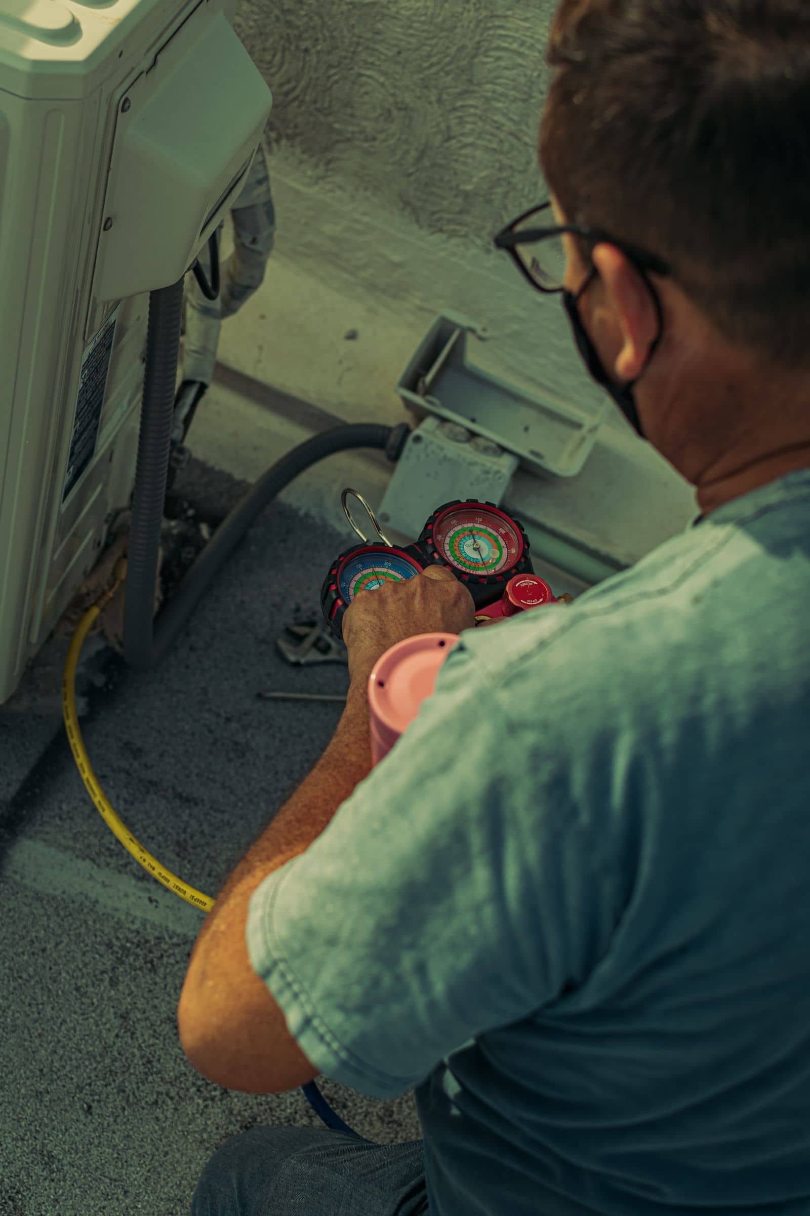 Technician kneeling at an outdoor condenser with manifold gauges connected, reading refrigerant pressures