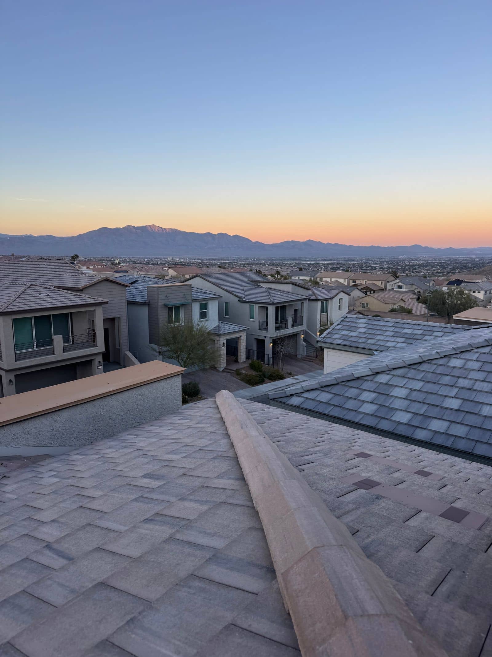 Las Vegas hillside neighborhood at twilight, rooftops stepping down toward the valley floor and mountains