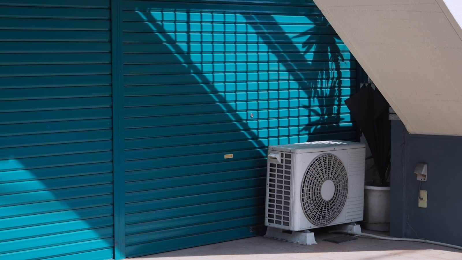 Heat pump condenser on a concrete pad in hard desert sunlight, palm shadow across a teal wall behind it